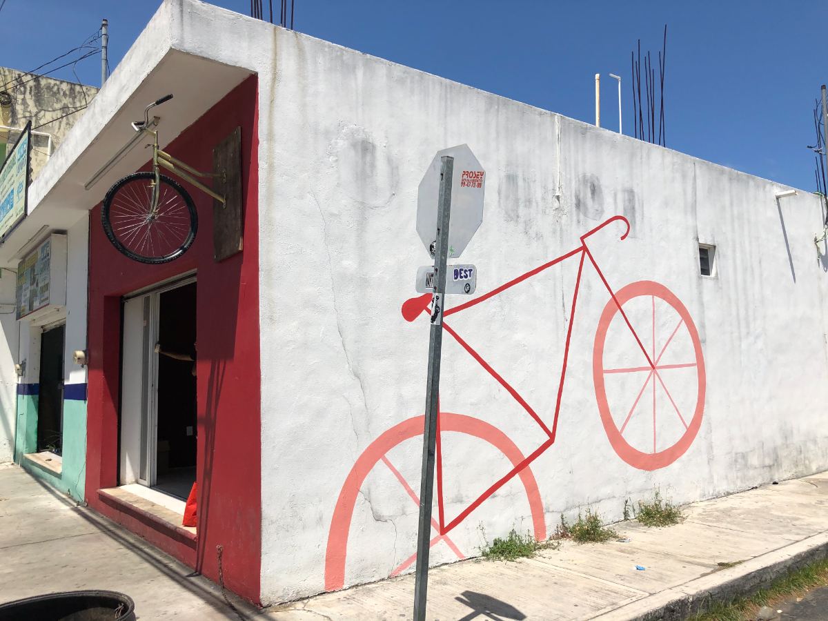 Red line-drawn bicycle painted across a white corner storefront wall — hand-painted mural in Playa del Carmen, Mexico by Scout Hill Co
